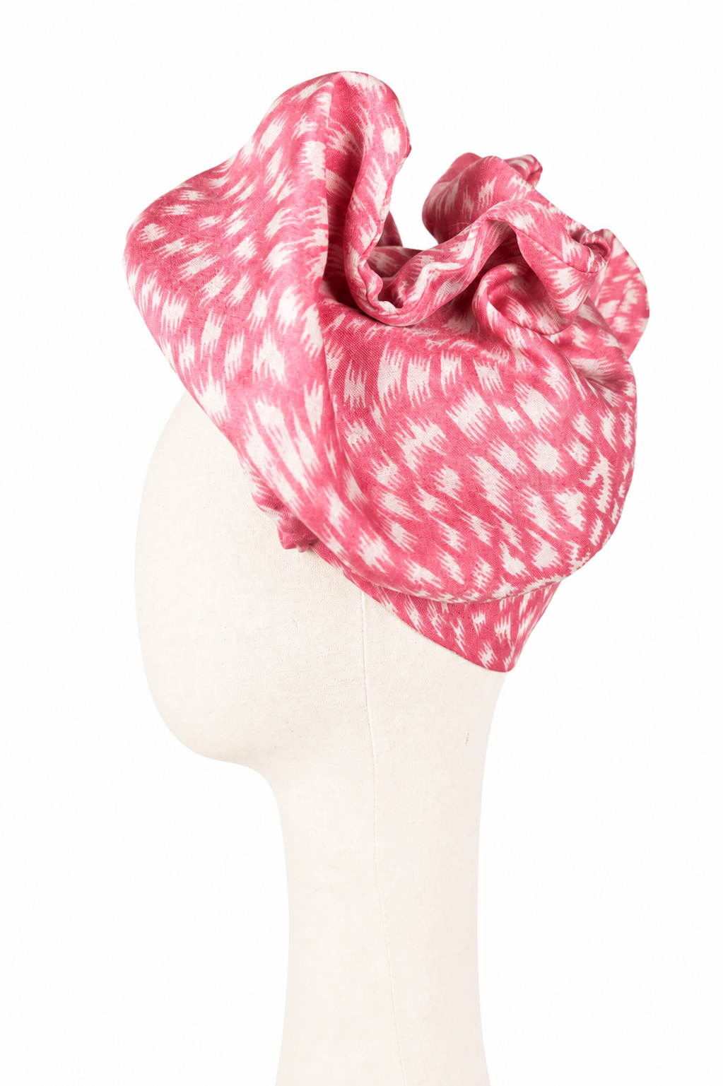 Lewy profil kremowego manekina w różowym turbanie Twist Long z pashminy i jedwabiu. Turban tworzy wysoką, przestrzenną formę z szerokich miękkich fałd i zawinięć. Malinoworóżowa tkanina ma biały, nieregularny nadruk przypominający postrzępione kreski i drobne cętki. Tło jest jasne i jednolite.