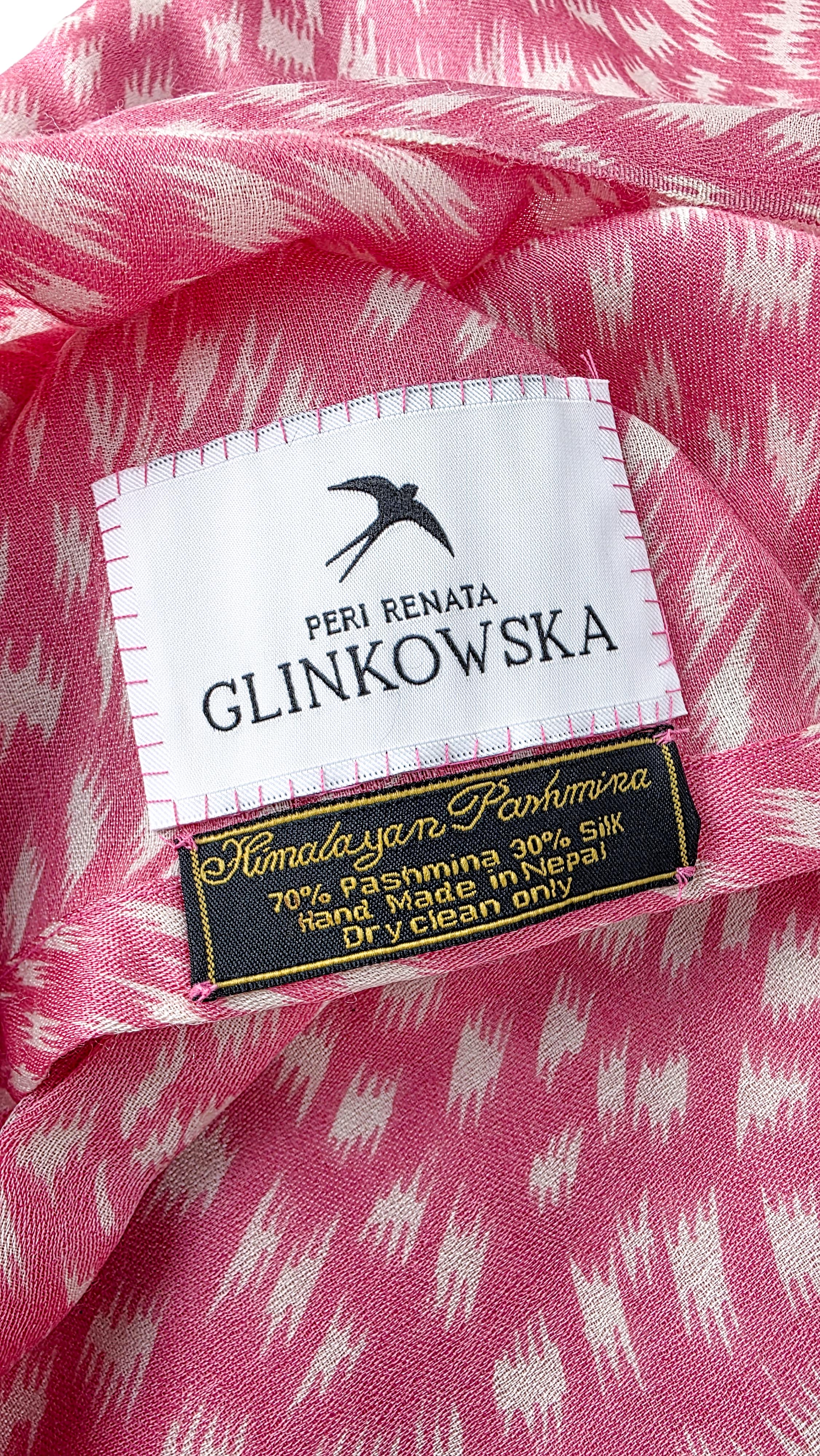 Zbliżenie wnętrza różowego turbanu z dwiema metkami. Górna jest biała z czarną jaskółką i napisem PERI RENATA GLINKOWSKA, przyszyta różową nicią. Pod nią czarna metka ze złotą obwódką i napisem Handmade yarn Pashmina oraz informacją 70% Pashmina, 30% Silk, Made in Nepal, Dry clean only.