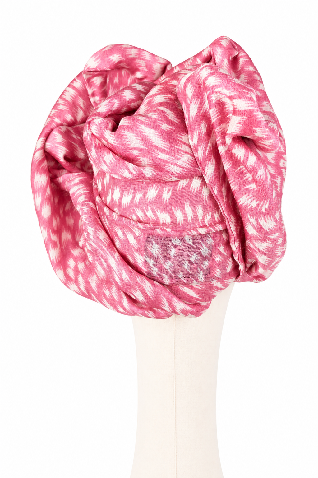 Tył różowego turbanu Twist Long z pashminy i jedwabiu na kremowym manekinie. Materiał tworzy warstwowe, asymetryczne pętle, rulony i miękkie fałdy, które nachodzą na siebie i budują rzeźbiarską formę. Malinoworóżową tkaninę pokrywa jasny, nieregularny nadruk w postrzępione kreski i plamy.