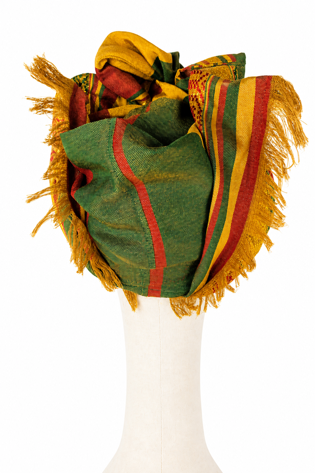 Widok od tyłu na turban Twist Long udrapowany na kremowym manekinie. Centralną część tworzy duża zielona płaszczyzna przecięta ceglastoczerwonym pasem. Po bokach materiał zawija się w przestrzenne fałdy z pasami zieleni, złota i czerwieni. Wokół obu boków i dolnej krawędzi widoczne są liczne złociste frędzle z oryginalnego etiopskiego szala.