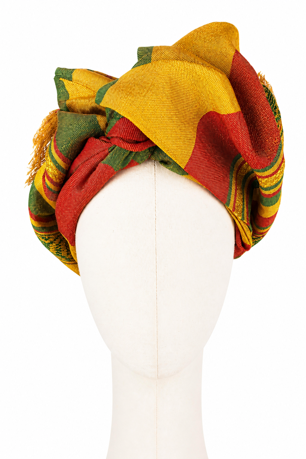 Turban damski Twist Long pokazany od przodu na kremowym manekinie bez rysów twarzy. Tkanina w kolorach zieleni, złocistej żółci i ceglastoczerwonego jest wysoko udrapowana w szerokie, asymetryczne fałdy. Nad czołem krzyżują się czerwone i zielone pasy, a duża złota fałda tworzy najwyższy punkt turbanu. Po bokach widoczne są tkane ornamenty i frędzle.