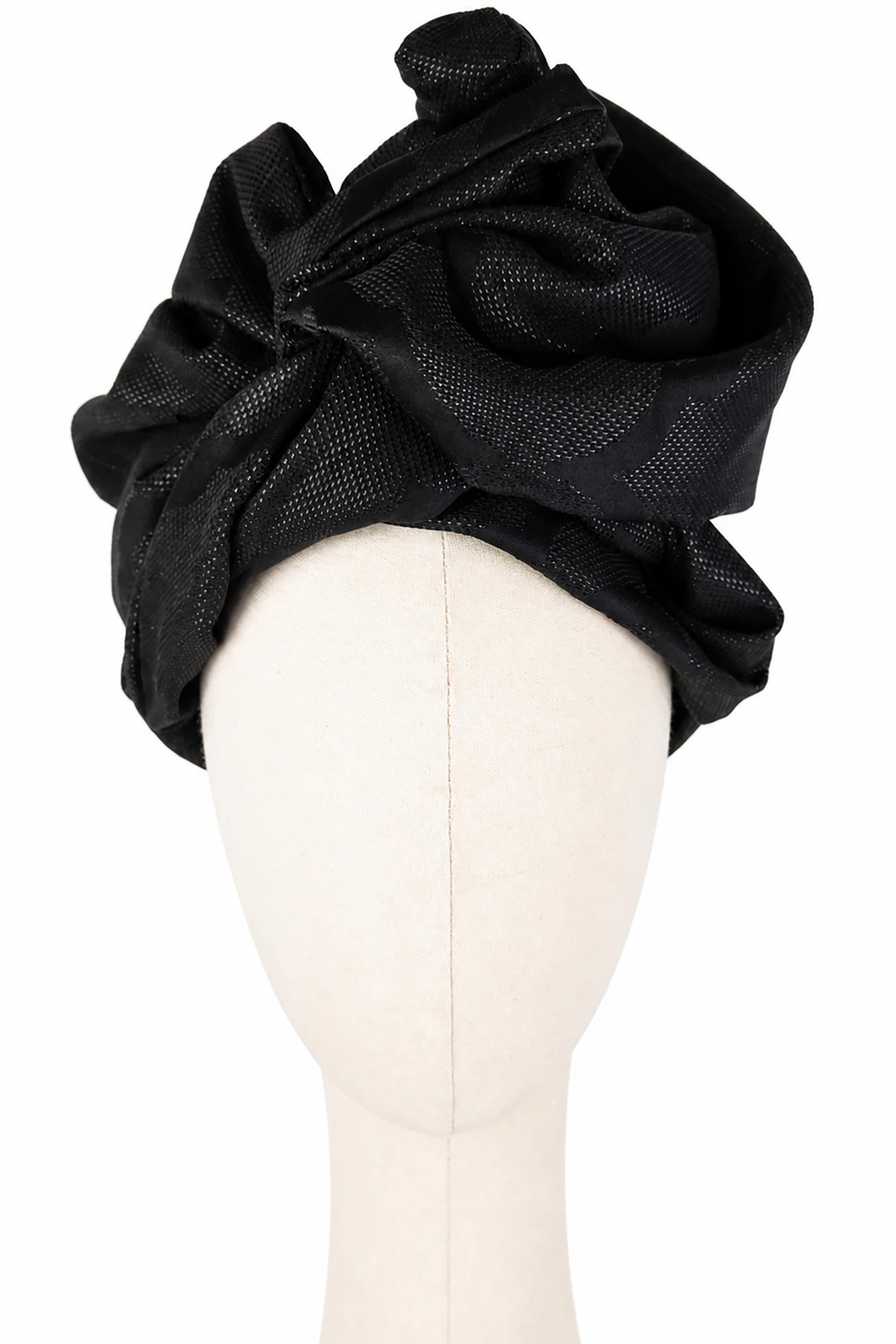 Czarny turban damski Twist Long pokazany od przodu na jasnym manekinie bez rysów twarzy. Materiał jest wysoko udrapowany nad czołem w szerokie, miękkie fałdy i asymetryczne zawinięcia. Na wypukłych częściach tkaniny widoczny jest subtelny połysk i duży ornament żakardowy, który miejscami pojawia się wyraźniej, a miejscami znika w matowej czerni.