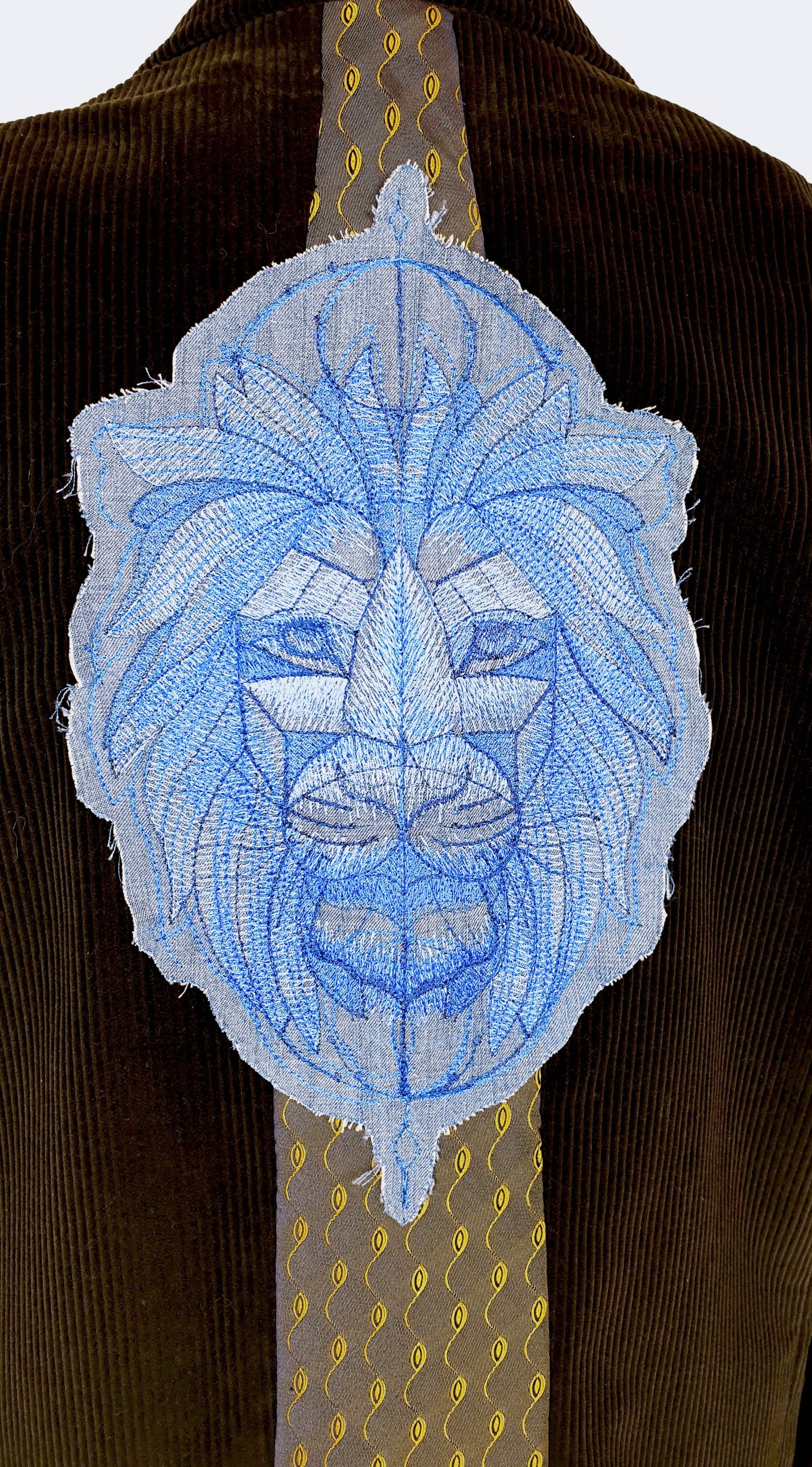 Gros plan : une tête de lion brodée en point serré dans plusieurs nuances de bleu sur un denim clair aux bords effilochés. On distingue les mèches géométriques de la crinière, les yeux et le museau. L'appliqué repose sur une cravate en soie gris-bleu à gouttes dorées, cousue sur le velours brun.