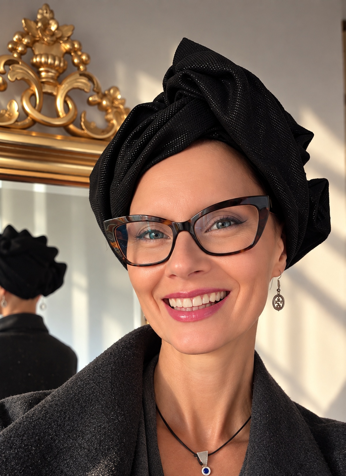Uśmiechnięta kobieta w czarnym turbanie damskim Twist Long z tkaniny żakardowej, pionowe ujęcie od przodu. Wysoka, przestrzenna forma z szerokich fałd obramowuje twarz, a na tkaninie połyskuje subtelny ornament. Kobieta nosi okulary w szylkretowych oprawkach, srebrne kolczyki i ciemnoszary płaszcz; w tle złocona rama lustra w ciepłym świetle.