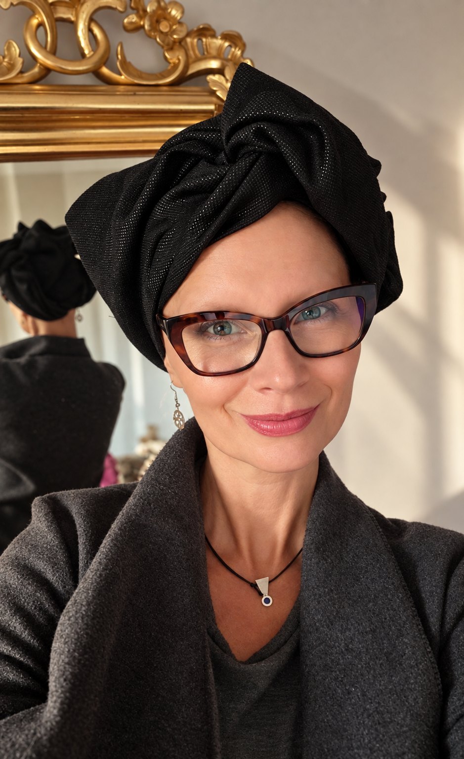 Kobieta w czarnym turbanie damskim Twist Long z tkaniny żakardowej, ujęcie z lekkim skrętem w lewo. Turban jest wysoko udrapowany nad czołem i tworzy z przodu duży, skręcony węzeł z szerokich fałd. Kobieta nosi okulary w szylkretowych oprawkach, srebrny wisior i ciemnoszary płaszcz; w tle złocona rama lustra i jasna ściana w ciepłym świetle.