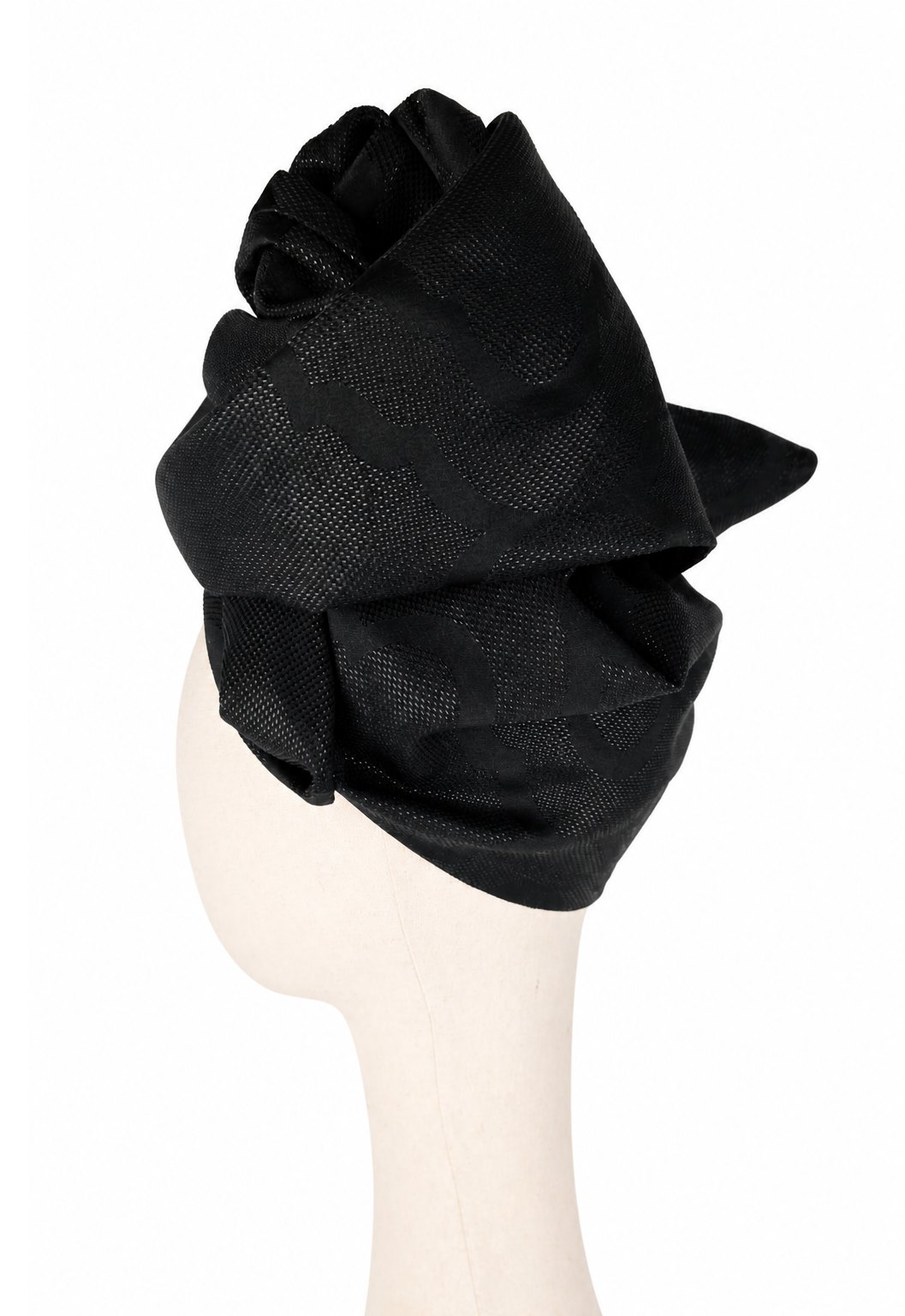 Lewy profil jasnego manekina w czarnym turbanie damskim Twist Long. Materiał jest udrapowany wysoko nad głową i tworzy kilka dużych, miękkich zawinięć. W żakardowej tkaninie widoczny jest ornamentowy wzór w czerni, ujawniający się dzięki różnicy między nićmi matowymi i lekko błyszczącymi. Forma turbanu jest elegancka i wyraźnie przestrzenna.
