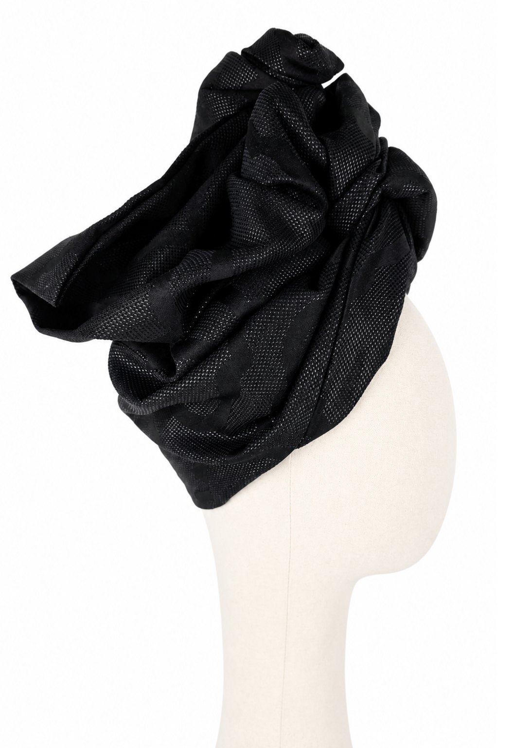 Prawy profil jasnego manekina w czarnym turbanie Twist Long. Drapowanie tworzy wysoką, przestrzenną konstrukcję z kilku szerokich fałd. Na bocznej powierzchni widoczny jest duży tkany ornament, którego błyszczące nici odbijają światło. Czerń przechodzi od głębokiej matowej do lekko połyskującej, podkreślając kształt turbanu.