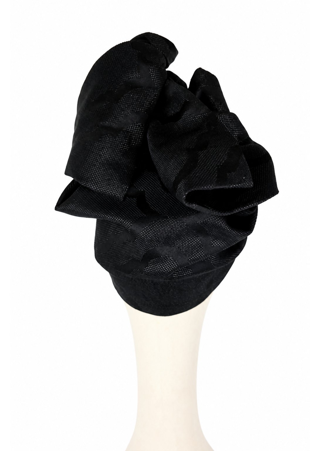 Widok od tyłu na czarny turban Twist Long udrapowany na jasnym manekinie. Tkanina układa się w szerokie zakładki i skręcone fałdy, tworząc zwartą, przestrzenną formę. W tylnej części widoczne są fragmenty dużego ornamentowego wzoru żakardowego. Subtelny połysk nici pojawia się głównie na krawędziach fałd, gdzie pada światło.