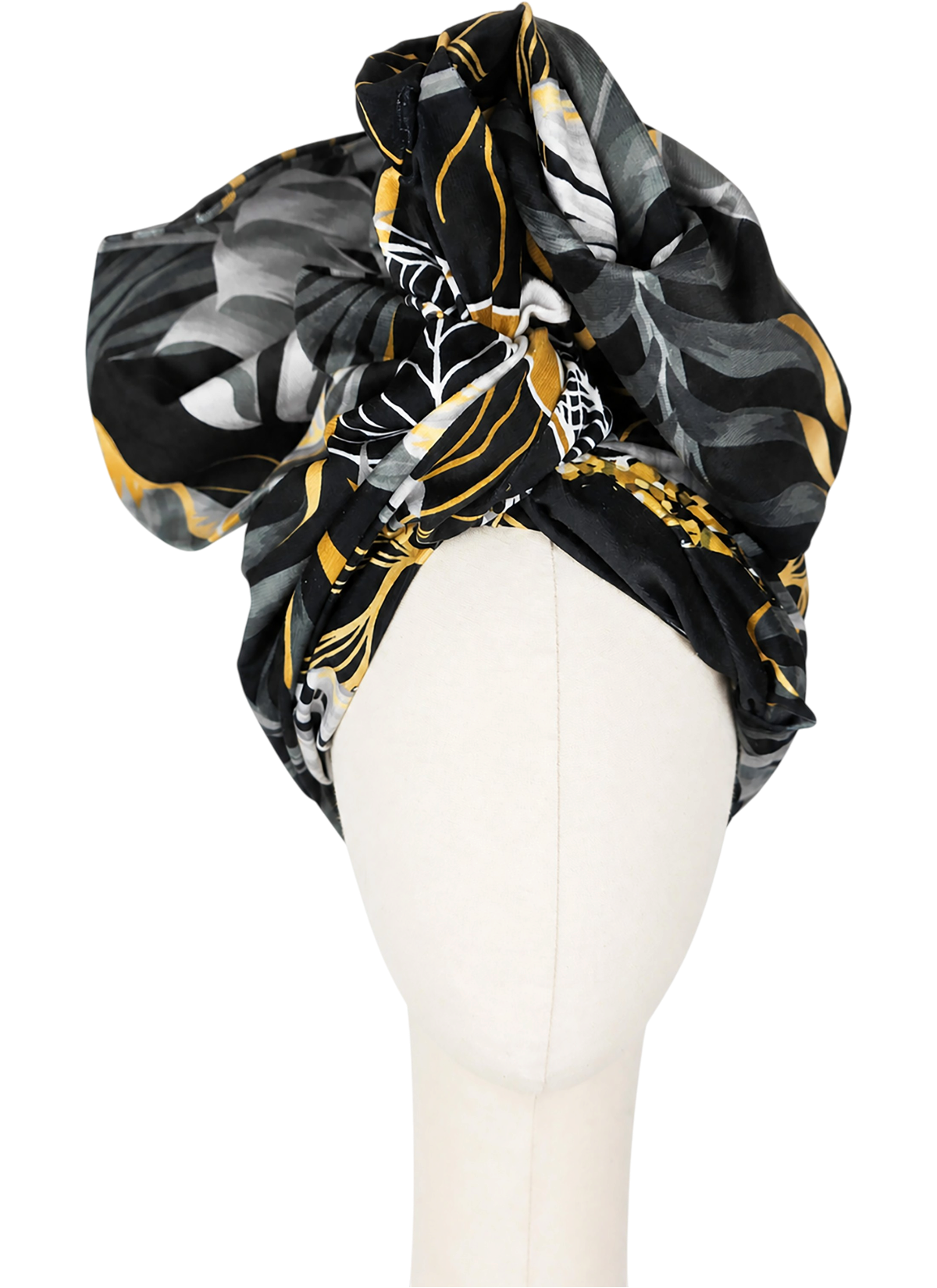 The women’s Twist Long turban is shown from the front on a light-coloured, featureless mannequin. The thicker fabric is styled high in broad, soft folds. Large white and golden-yellow tropical leaves stand out against the black and grey background. The pattern shifts across the draping, so each section reveals a different arrangement of leaves.