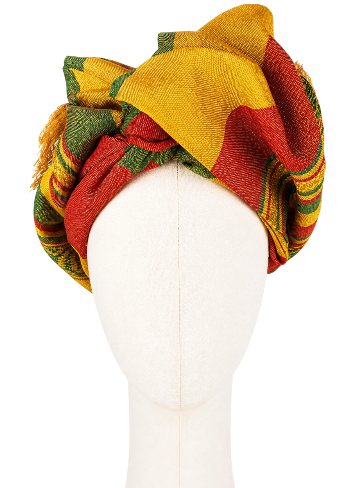 Women's Twist Long turban shown from the front on a cream-colored faceless mannequin. The fabric in shades of green, golden yellow, and brick red is draped high into wide, asymmetrical folds. Red and green bands cross above the forehead, while a large golden fold forms the highest point of the turban. Woven ornamental patterns and fringes are visible on the sides.