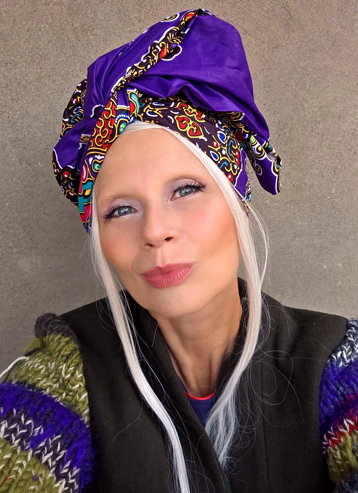 Woman wearing a purple African print turban tied high with a sculptural side bow, against a gray concrete wall.
