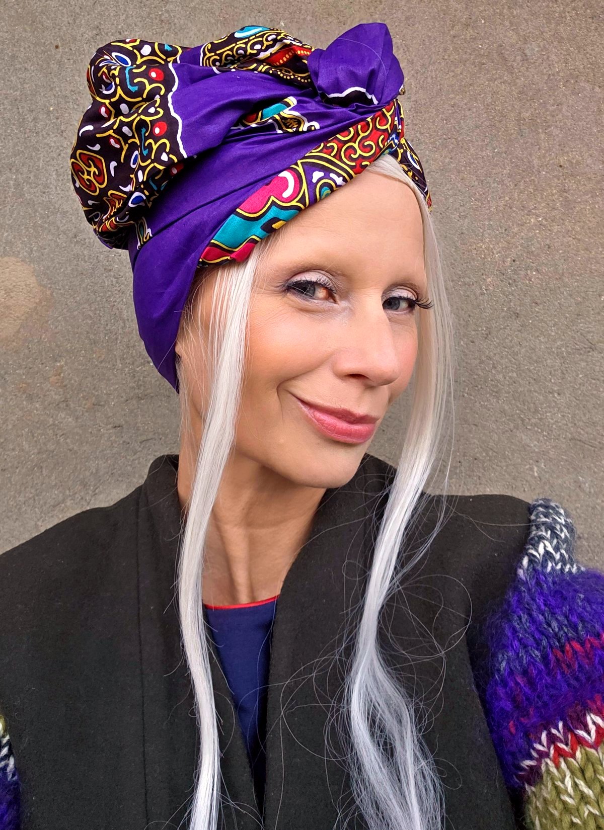 Woman with long white hair wearing a purple ornamental print turban tied with a small knot on top.