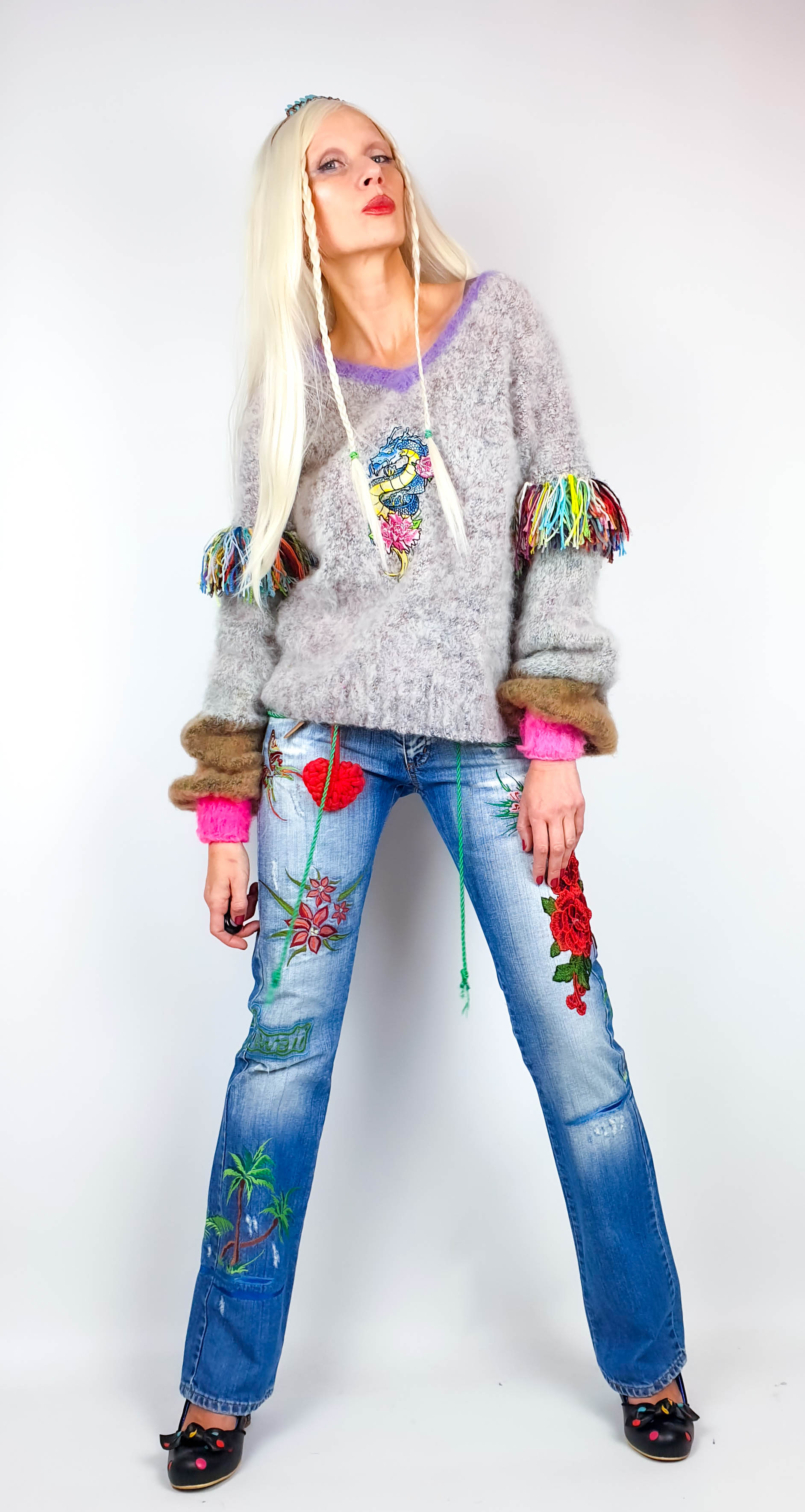 Full-length view of a woman in a fluffy oversize sweater with dragon embroidery, rainbow fringe and neon pink cuffs.
